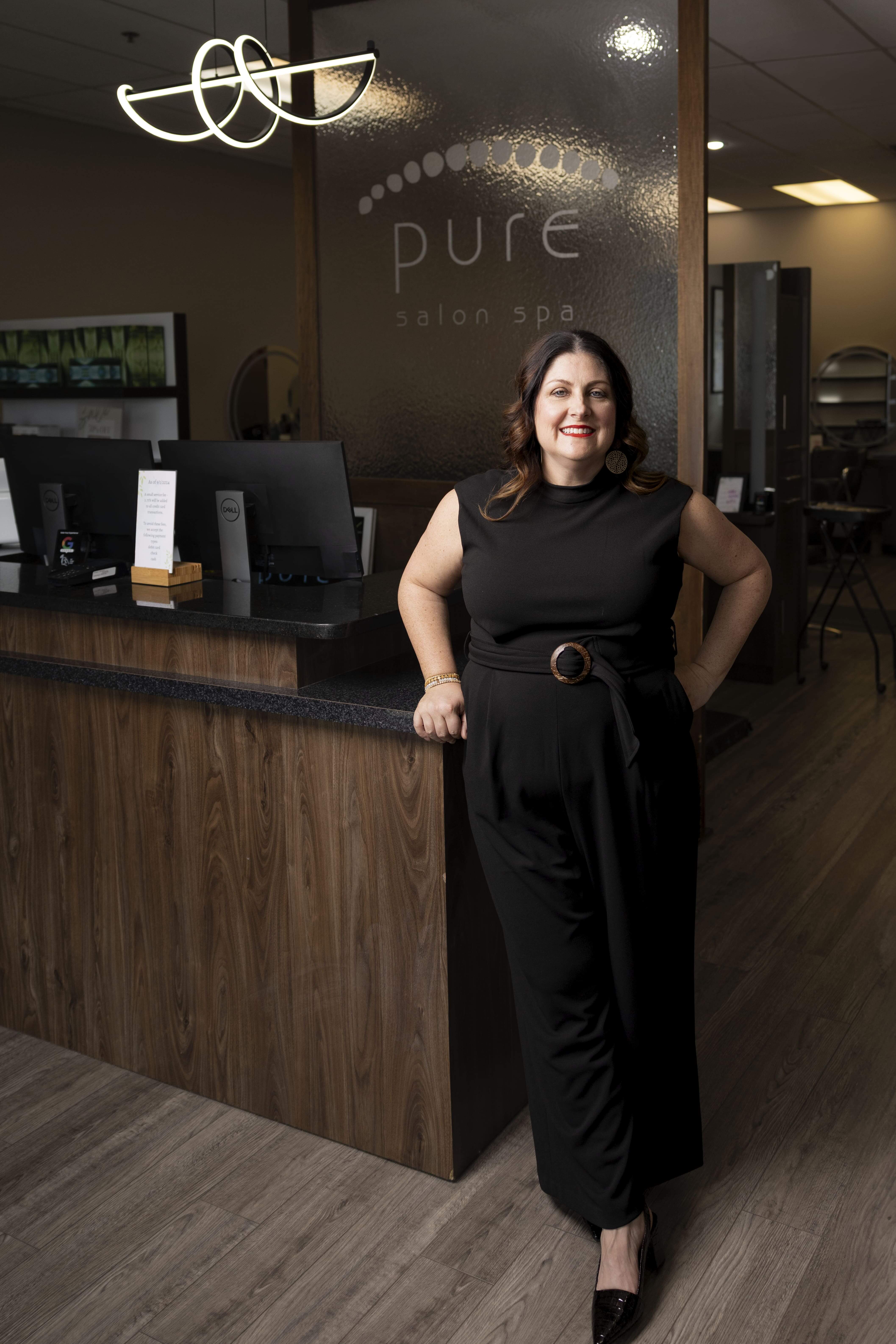 A woman in a black dress stands at the reception desk of Pure Salon Spa, smiling at the camera. The salon interior is modern with wood accents. - Pure Salon Spa, KY