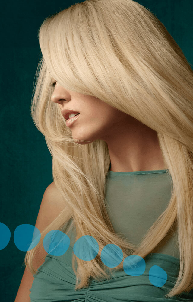 A woman with long, straight, platinum blonde hair colored with Aveda professional hair color faces sideways, partially covering her face, wearing a sleeveless green top against a dark teal background with blue circles. - Pure Salon Spa, KY