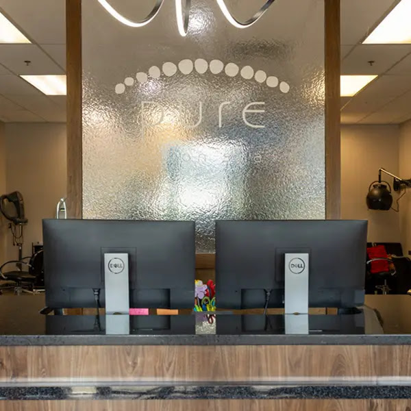 Front desk with two Dell monitors in a modern salon reception area, featuring a textured glass wall with the word "PURE" displayed behind the desk. - Pure Salon Spa, KY