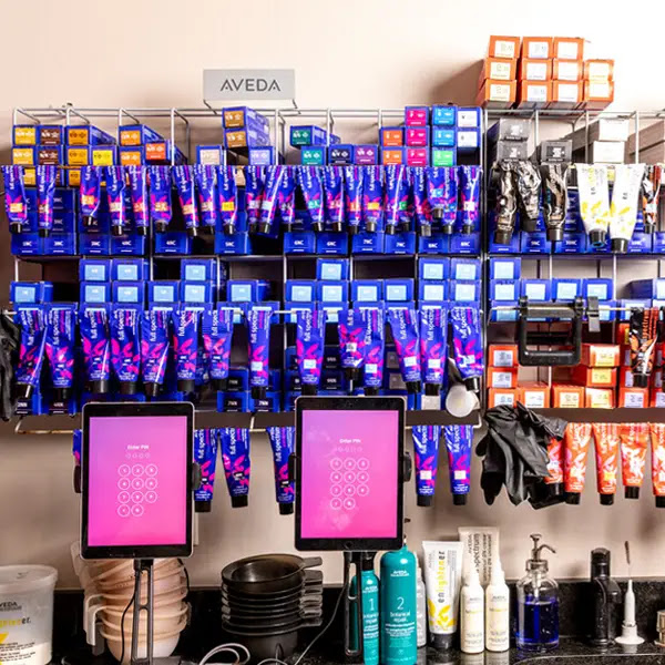 Shelves filled with colorful hair dye tubes and boxes, hair products, gloves, and tablets with pink screens on a salon counter. - Pure Salon Spa, KY