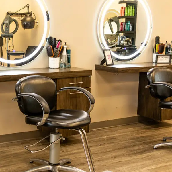 Two empty salon chairs sit in front of round mirrors with illuminated edges, with hair care products and brushes arranged on wooden counters. - Pure Salon Spa, KY