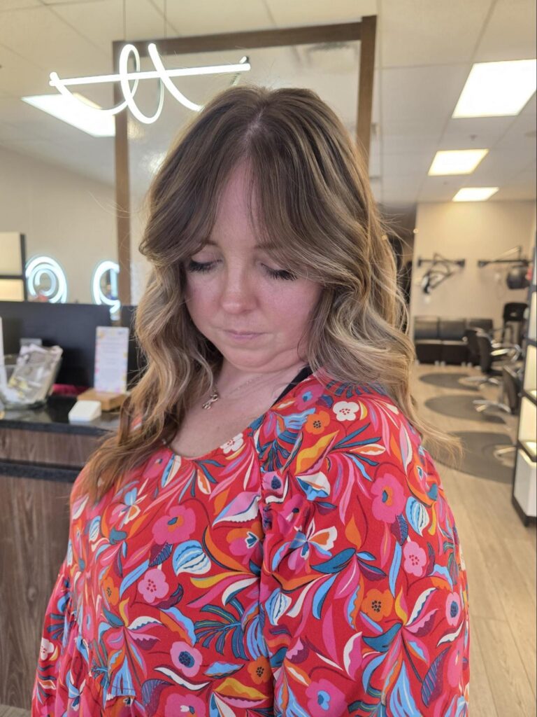 A woman with wavy, highlighted hair from the Bloom & Blend Collection wears a colorful floral top and looks down, standing in a modern Louisville KY hair salon. - Pure Salon Spa, KY