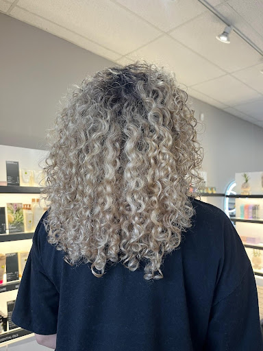 Person with shoulder-length, blonde curly hair seen from behind, standing in a modern, well-lit store in Louisville KY, surrounded by shelves of products—showcasing styles inspired by the Bloom & Blend Collection and Spring 2026 hair trends. - Pure Salon Spa, KY