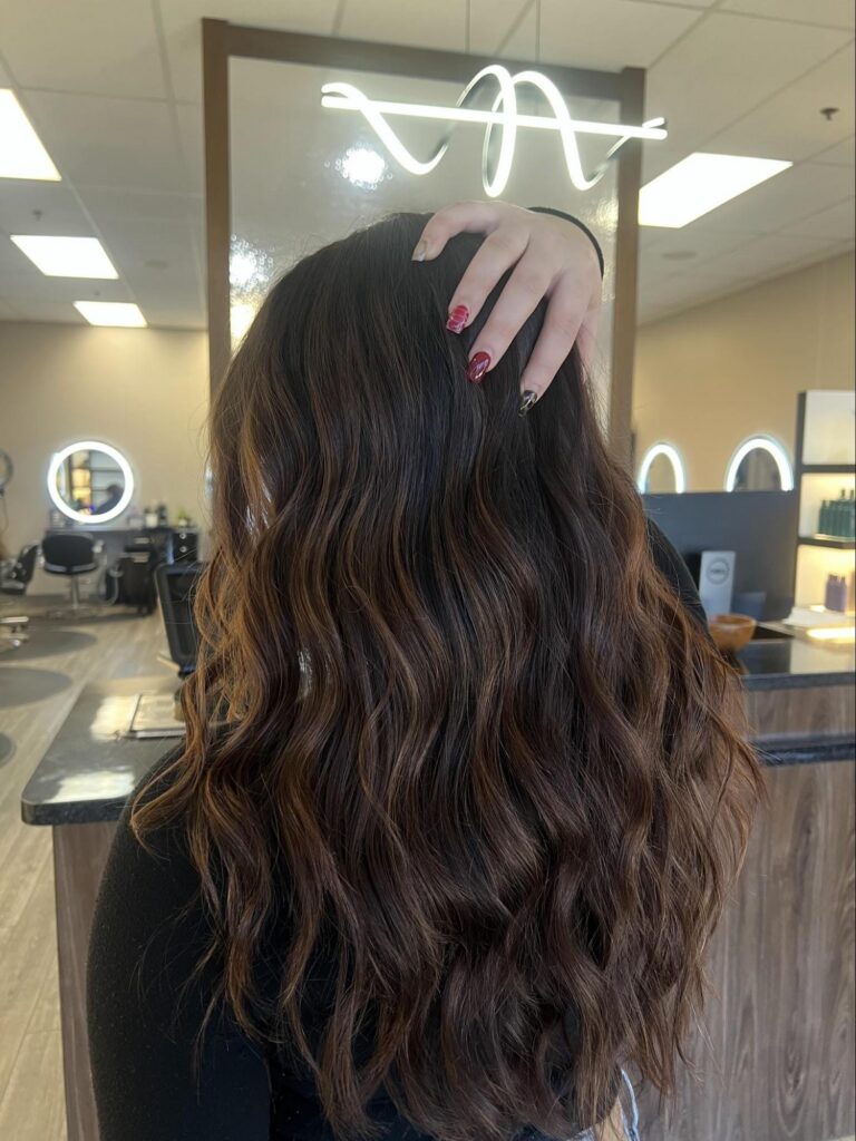 Showcasing long, wavy brown hair from the Bloom & Blend Collection, a person stands in a Louisville KY salon, facing away from the camera with one hand atop their head—embracing spring 2026 hair trends. - Pure Salon Spa, KY