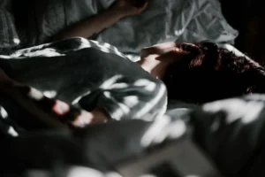 Person lying on a bed in a dimly lit room, with light and shadow patterns across their body and bedding—a quiet glimpse into what depression looks like. - Pure Salon Spa, KY