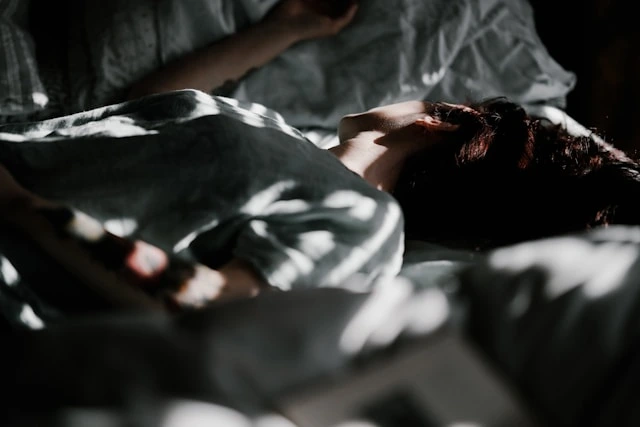 Person lying on a bed in a dimly lit room, with light and shadow patterns across their body and bedding—a quiet glimpse into what depression looks like. - Pure Salon Spa, KY